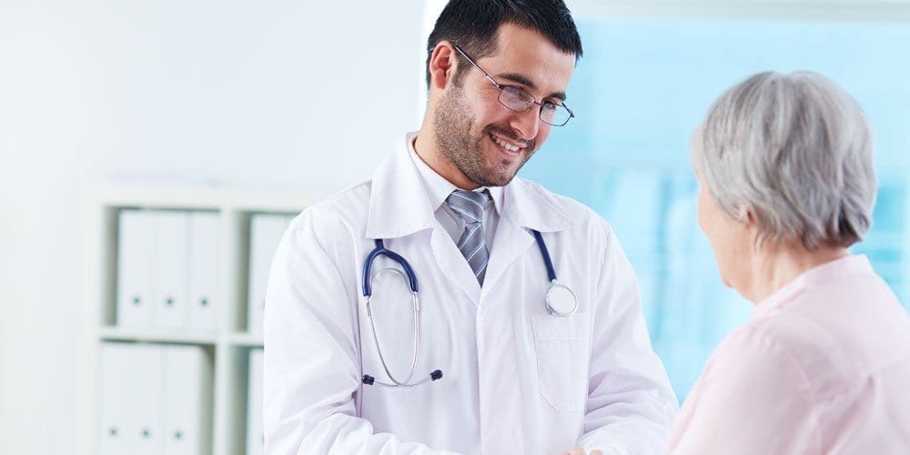 Male doctor smiling and talking with an elderly woman wearing a pink shirt, showing compassionate patient care.