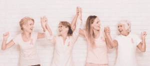 Four women wearing pink shirts with breast cancer awareness ribbons, standing together and holding hands in solidarity.